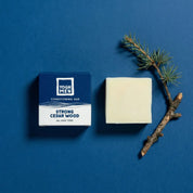 yoghmen conditioner bar for strong hair next to its packaging and cedar wood branch on a blue background